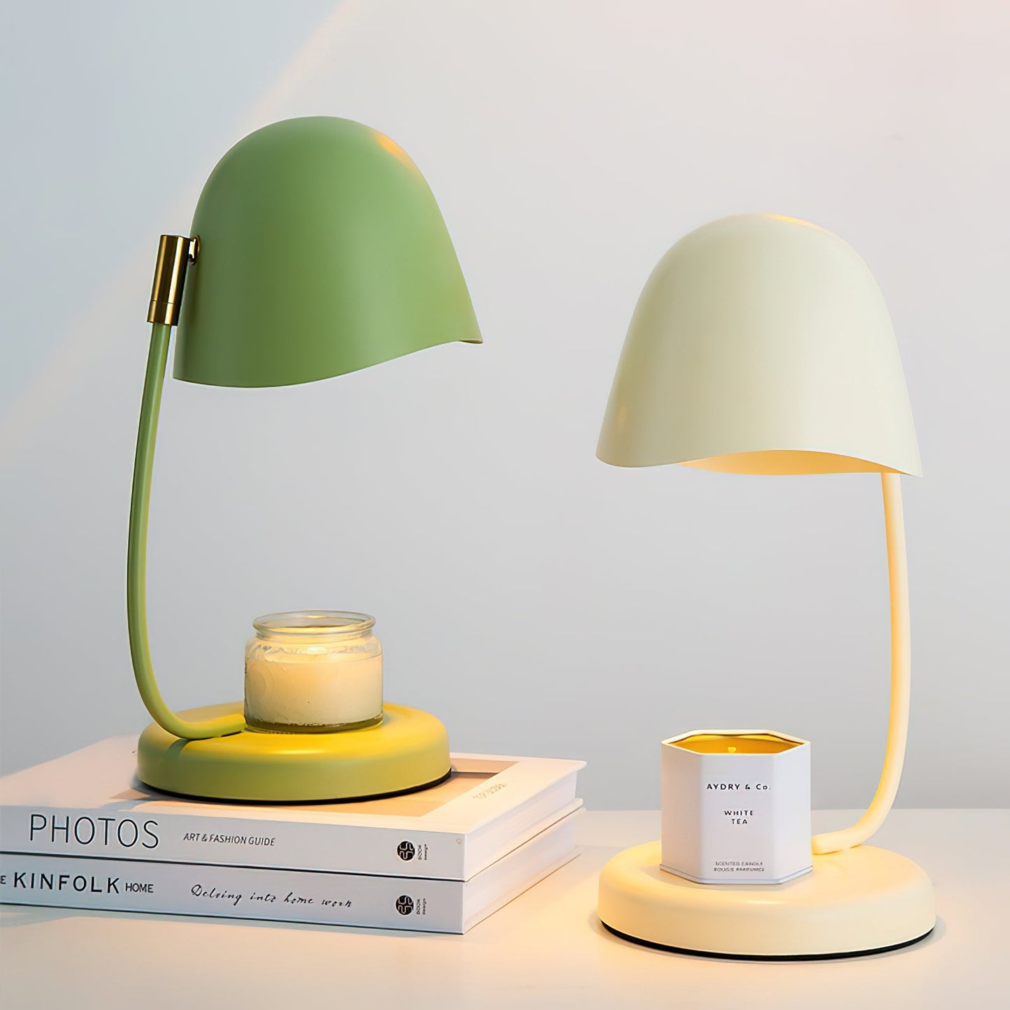 Two candle warmer lamps, one matcha green and one cream, on a surface with books and a candle.