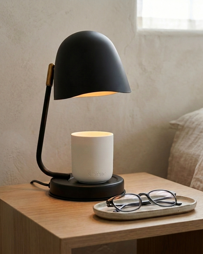 A black Solene candle warmer lamp with a white ceramic candle sits on a light oak bedside table, with glasses on a tray.